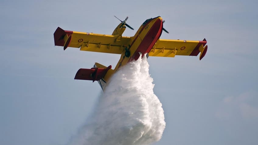 Ein gelb-rotes Löschflugzeug von unten, das eine große Wasserlast abwirft.