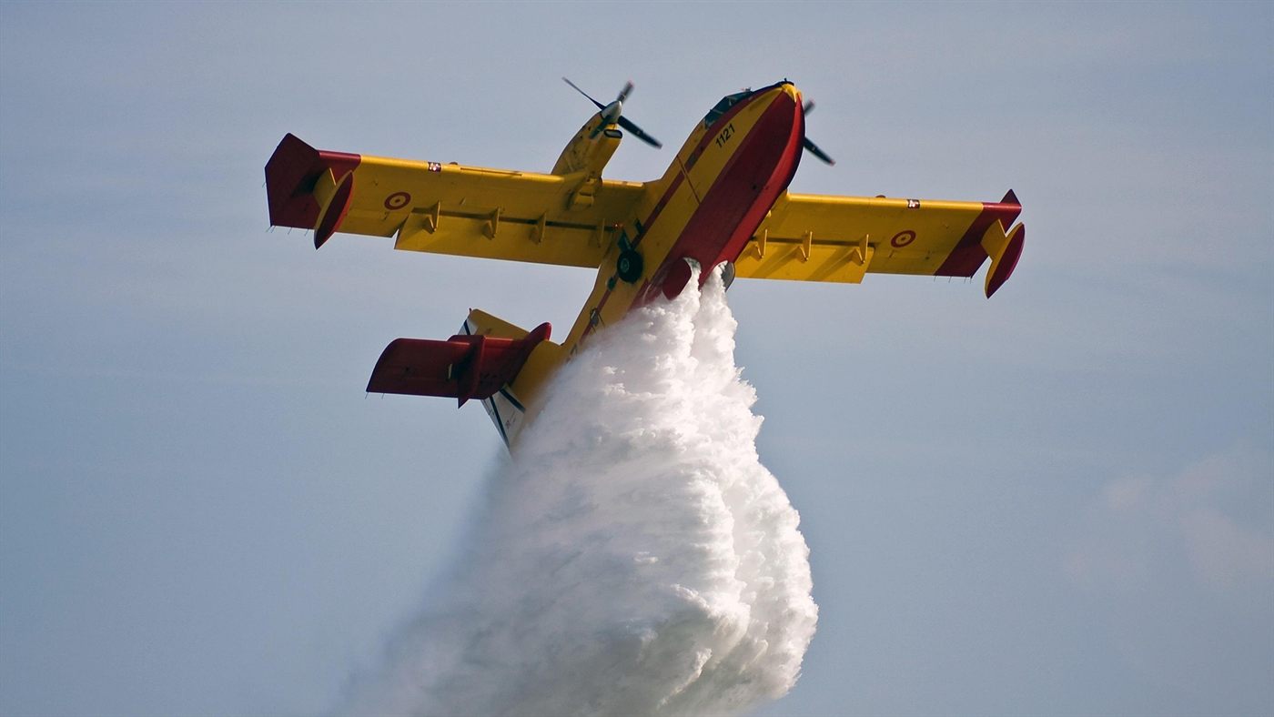 Ein gelb-rotes Löschflugzeug von unten, das eine große Wasserlast abwirft.