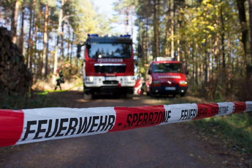 Ein Löschfahrzeug und ein kleineres Fahrzeug stehen auf einem sandigen Waldweg,
                davor ein Absperrband mit der Aufschrift Feuerwehr Sperrzone.