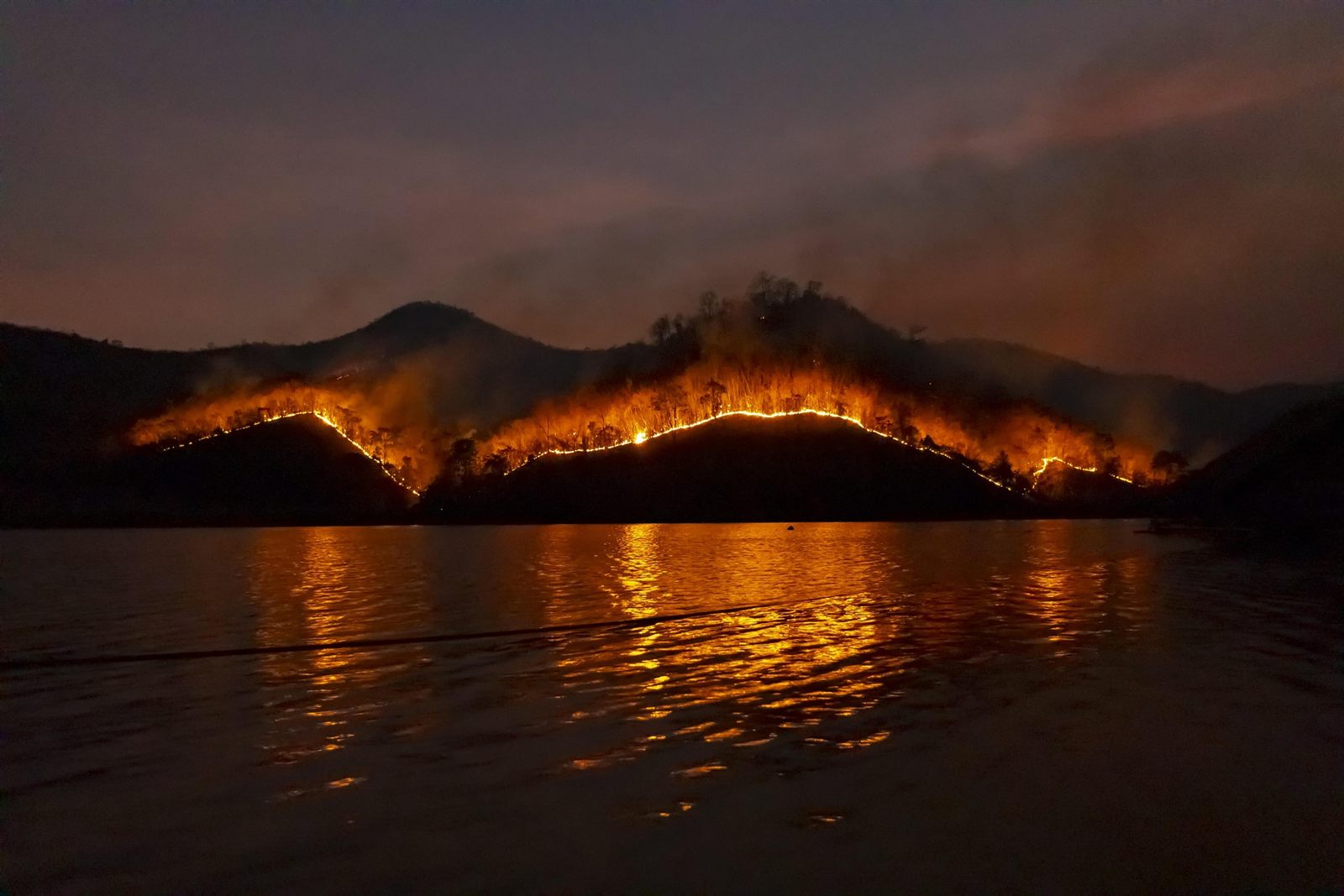 Ein Feuer brennt in der Dämmerung entlang eines bewaldeten Höhenzugs, sein Licht
                spiegelt sich im Wasser davor.