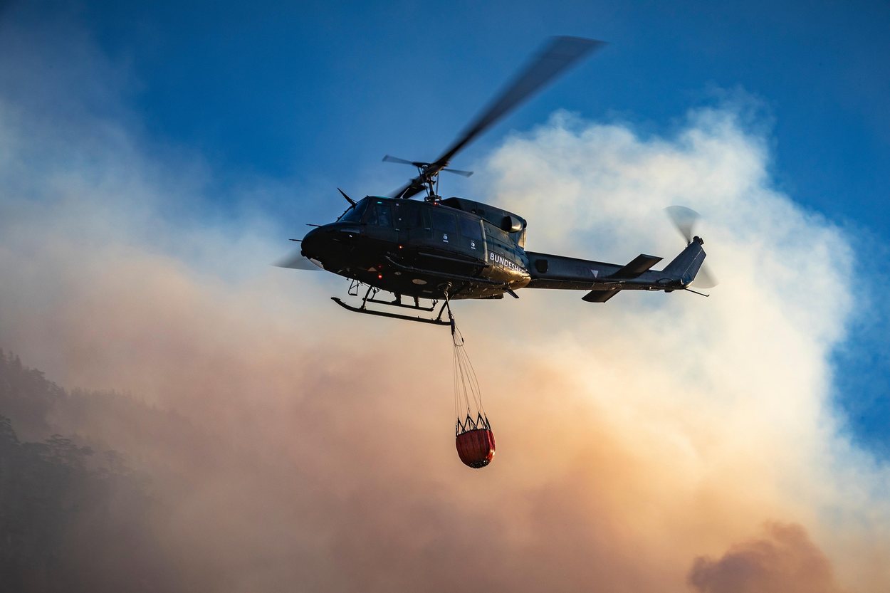 Ein Militärhubschrauber trägt einen Löschwasserbehälter an langer Leine über Rauch
                und einem bewaldeten Hang.